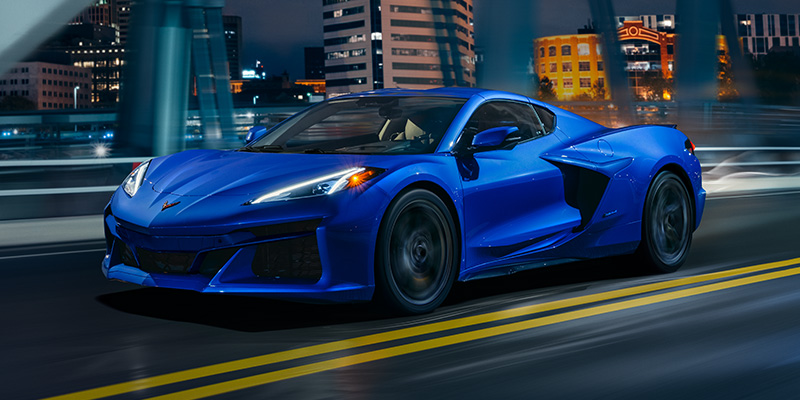 A blue 2026 Chevrolet Corvette sports car driving on a bridge at night.